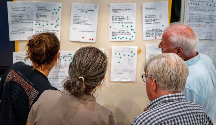 Teilnehmerinnen und Teilnehmer des Klima-Bürgerrats in Pinneberg betrachten und diskutieren die an einer Pinnwand festgehaltenen Arbeitsergebnisse. Auf mehreren Blättern sind Vorschläge und Bewertungen gesammelt und mit Klebepunkten gewichtet worden.